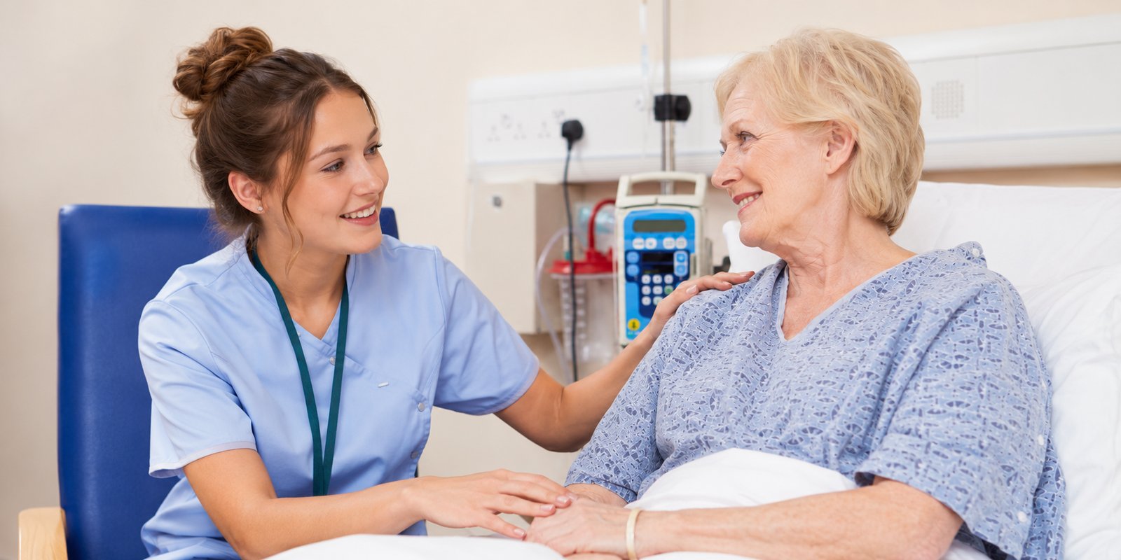 A smiling medical assistant in blue scrubs sits at an elderly patient's bedside, one hand on her shoulder and the other on her hand.