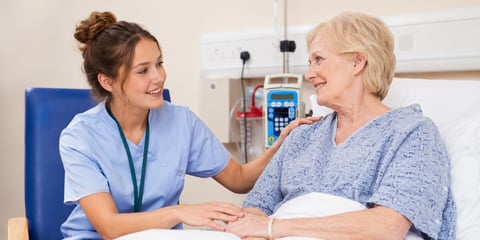 A smiling medical assistant in blue scrubs sits at an elderly patient's bedside, one hand on her shoulder and the other on her hand.